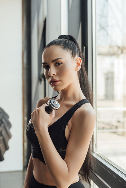 Beautiful sportswoman holding dumbbell and looking at camera near window in gym - Foto, afbeelding