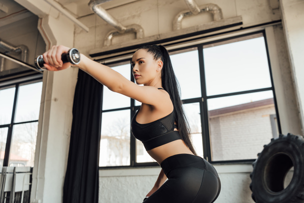 Sportswoman holding dumbbell with outstretched hand and doing squat in gym - Photo, Image