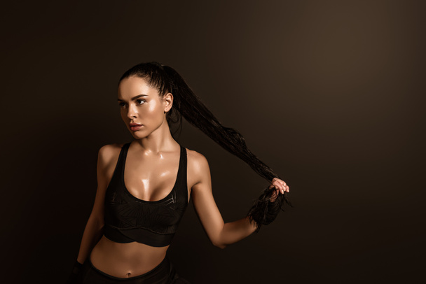 Sexy brünette Sportlerin berührt Haare und schaut isoliert auf schwarz weg - Foto, Bild