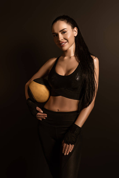 Brunette sportswoman with ball looking at camera and smiling isolated on black - Foto, Imagen