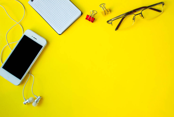 a phone with a blank screen on a yellow background, white vacuum headphones, Notepad, paper clips, and glasses. mock up. banner with space for text. business concept - Foto, Imagen