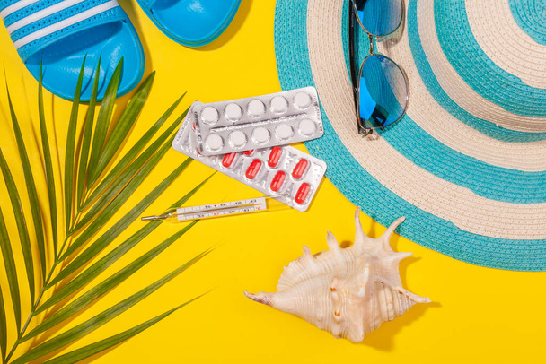 Top view on white and red pills in blister pack, blue hat, blue flip-flops, sunglasses, seashell, palm leaf on yellow background. Concept of beach holiday, sea tour, warm sunny summer. Acclimatization - Φωτογραφία, εικόνα