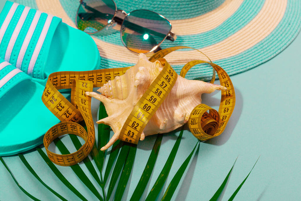 Vista dall'alto di cappello a righe, occhiali da sole, nastro di misura con una conchiglia e un ramo di palma su uno sfondo blu turchese. Concetto di perdere peso per una vacanza in mare. Spazio pubblicitario - Foto, immagini