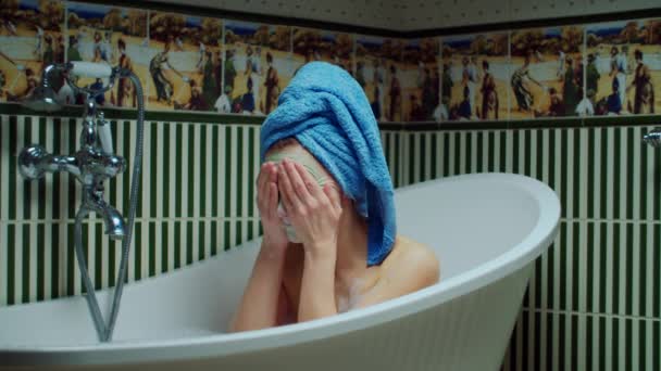Young 30s woman washing off facial mask in bathtub with foam at green bathroom at home. Woman with towel on head making bath procedures. Close up - Footage, Video
