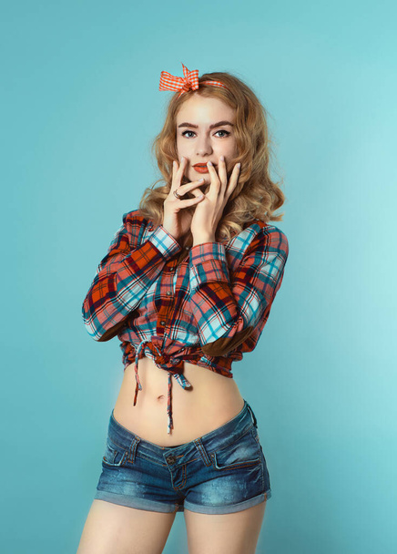 Pin-up girl aux cheveux blonds bouclés avec arc à carreaux rouges et chemise portant un short en jeans. - Photo, image