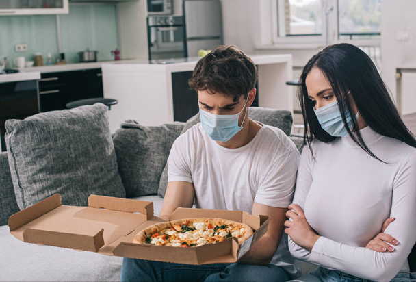 άρρωστο ζευγάρι με ιατρικές μάσκες κοιτάζοντας πίτσα στο σπίτι κατά τη διάρκεια της αυτο-απομόνωσης - Φωτογραφία, εικόνα