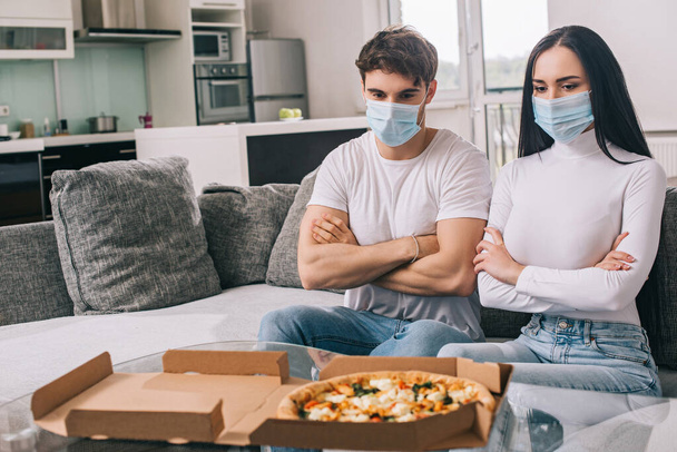 sad ill couple in medical masks with crossed arms looking at pizza at home during self isolation - Foto, Bild