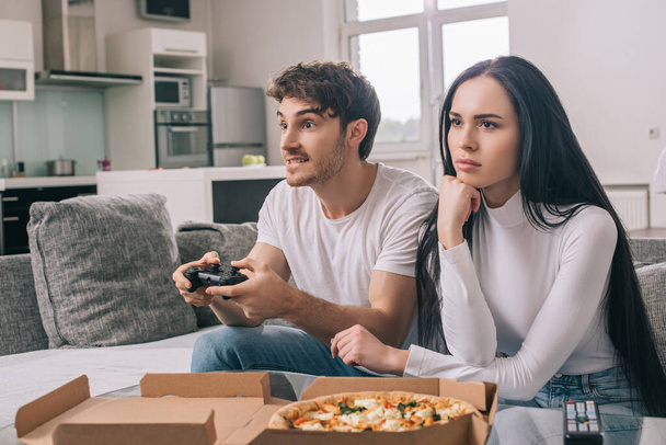 KYIV, UKRAINE - APRIL 16, 2020: sad girl sitting near boyfriend playing video game with joystick during self isolation at home with pizza - Photo, image