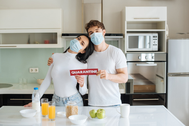 ζευγάρι με ιατρικές μάσκες που παίρνουν πρωινό και κρατούν το σήμα καραντίνας στο σπίτι κατά τη διάρκεια της αυτοαπομόνωσης - Φωτογραφία, εικόνα