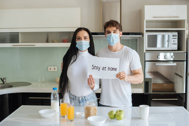 άρρωστο ζευγάρι με ιατρικές μάσκες που παίρνουν πρωινό και κρατούν Μείνετε στο σπίτι σημάδι στο σπίτι κατά τη διάρκεια της αυτοαπομόνωσης - Φωτογραφία, εικόνα