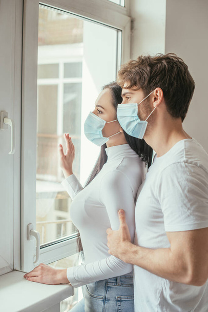 beautiful couple in medical masks looking through window during self isolation - Foto, Imagen