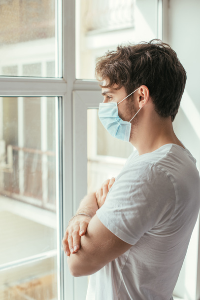 mladý smutný muž v lékařské masce se zkříženýma rukama dívá oknem během vlastní izolace  - Fotografie, Obrázek