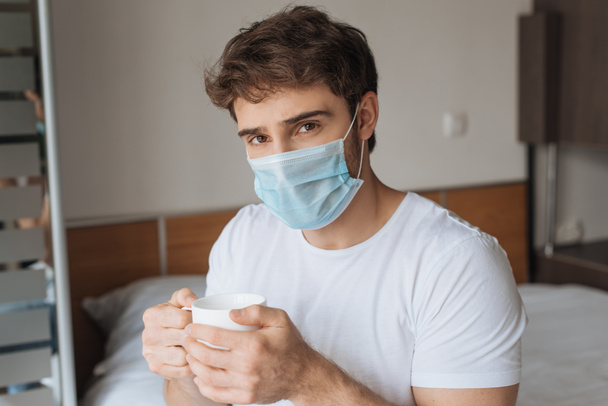 junger kranker Mann in medizinischer Maske hält Tasse mit Heißgetränk während der Selbstisolierung auf dem Bett - Foto, Bild