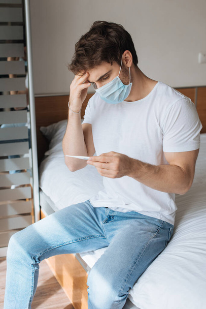 κουρασμένος άρρωστος άνθρωπος με ιατρική μάσκα κοιτάζοντας το θερμόμετρο κατά τη διάρκεια της αυτο-απομόνωσης - Φωτογραφία, εικόνα