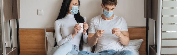 Worried couple in medical masks holding cup and thermometer during self isolation, panoramic crop - Foto, afbeelding