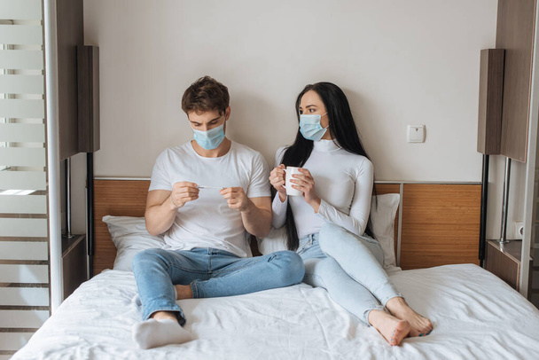 Huolestunut pari lääketieteellisissä naamioissa tilalla kuppi ja lämpömittari makuuhuoneessa aikana itsensä eristäminen
 - Valokuva, kuva