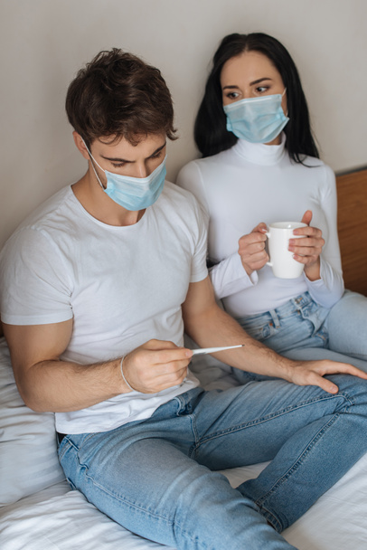 άρρωστο ζευγάρι με ιατρικές μάσκες κρατώντας κύπελλο και θερμόμετρο στο κρεβάτι κατά τη διάρκεια της αυτο-απομόνωσης - Φωτογραφία, εικόνα