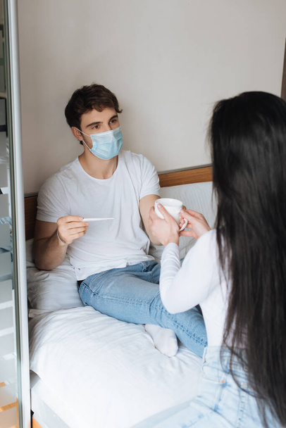 femme donnant tasse de boisson chaude à l'homme malade dans un masque médical avec thermomètre au lit pendant l'isolement personnel
 - Photo, image