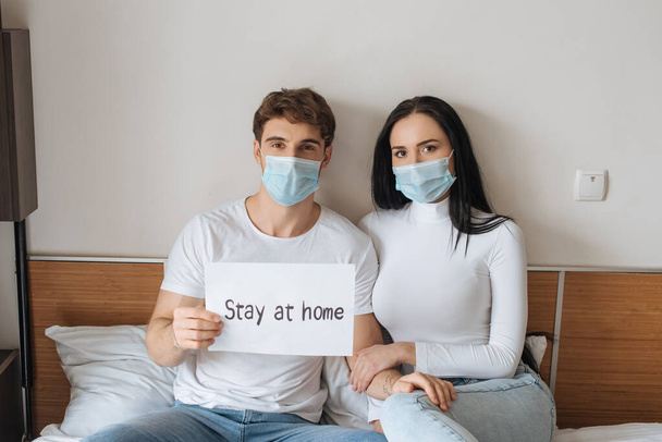 Krankes Paar in medizinischer Maske hält während der Selbstisolierung zu Hause Schild im Bett - Foto, Bild