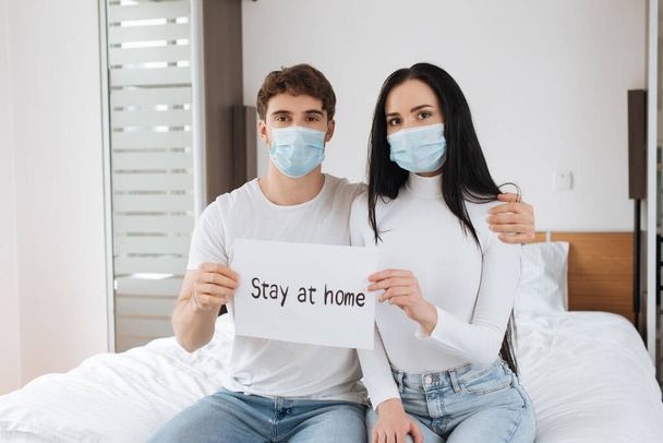 άρρωστο ζευγάρι με ιατρικές μάσκες που κρατούν Μείνετε στο σπίτι σημάδι στο σπίτι κατά τη διάρκεια της αυτο-απομόνωσης - Φωτογραφία, εικόνα