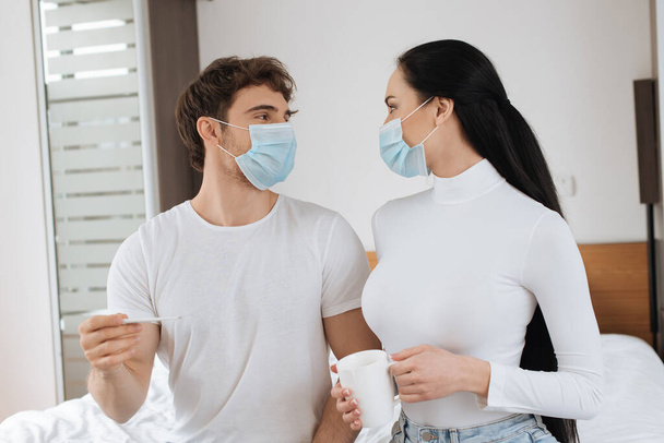 Krankes Paar in medizinischer Maske hält Becher und Thermometer in der Hand und schaut sich während der Selbstisolierung an - Foto, Bild