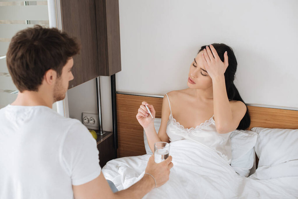 Mann schenkt kranker Freundin während Selbstisolierung ein Glas Wasser mit Thermometer im Bett  - Foto, Bild