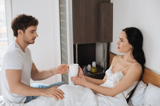boyfriend giving cup of hot drink to ill girlfriend in bed during self isolation  - 写真・画像