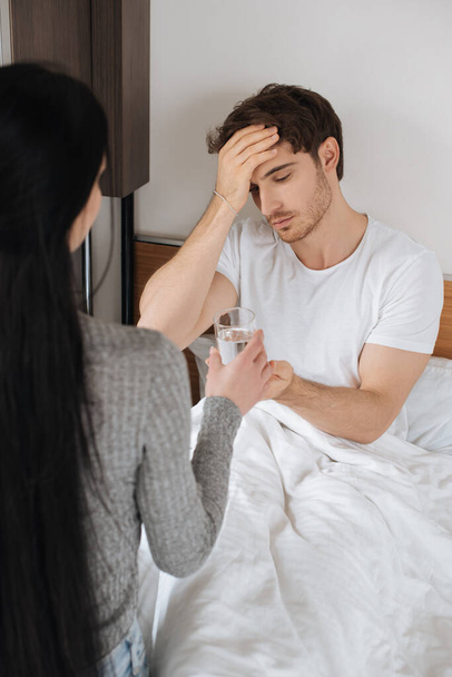 Frau gibt müdem Mann mit Kopfschmerzen während der Selbstisolierung ein Glas Wasser im Bett  - Foto, Bild