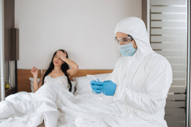 mannelijke arts in beschermend pak en medisch masker met behulp van smartphone in de slaapkamer met zieke vrouw  - Foto, afbeelding