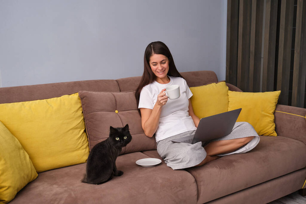 A brunette woman is sitting on the brown sofa with yellow cushions in the living room, wearing pyjamas, drinking tea, surfing the net on her laptop, looking at a black fluffy cat, happy to be at home - Фото, изображение