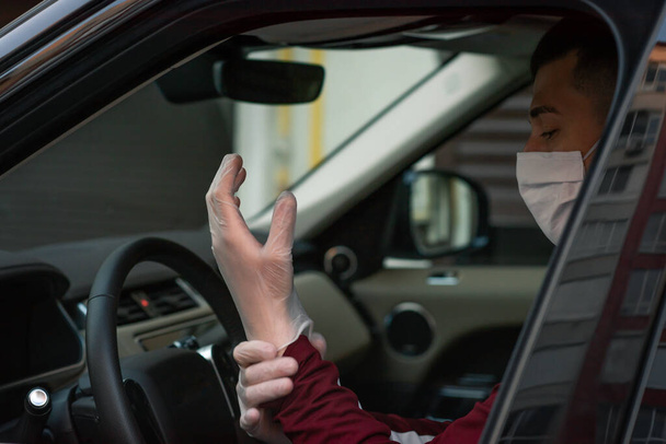 um homem dirigindo em máscara médica em carro coloca luvas médicas em um durante uma epidemia, um motorista de táxi em uma máscara, proteção contra o vírus. Condutor em carro preto. coronavírus, infecção, quarentena, covid-19
 - Foto, Imagem