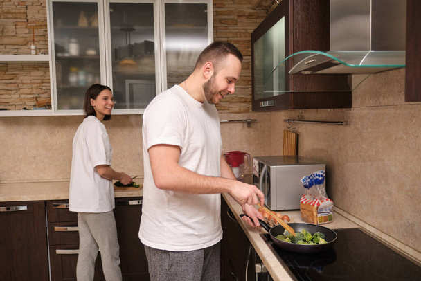 Happy young couple preparing breakfast, man fries broccoli, scrambled eggs. Girl slices fresh vegetables. On self-isolation during the coronovirus pandemic covid-19. - Photo, Image