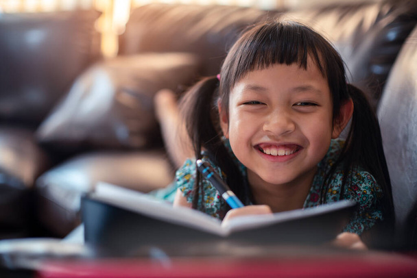 Ευτυχισμένο κοριτσάκι ξαπλωμένο στον καναπέ με μάθηση από φορητό υπολογιστή και γράφοντας ένα βιβλίο στο σπίτι, Κοινωνική απόσταση κατά τη διάρκεια της καραντίνας, Online εκπαιδευτική έννοια - Φωτογραφία, εικόνα