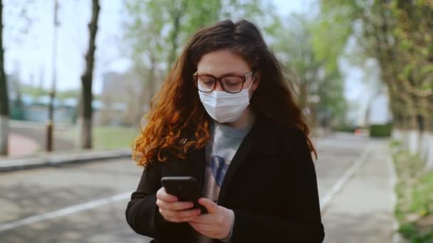 Nuori tyttö lääketieteellisessä naamiossa käyttää älypuhelinta puistossa. 4 k.
 - Materiaali, video