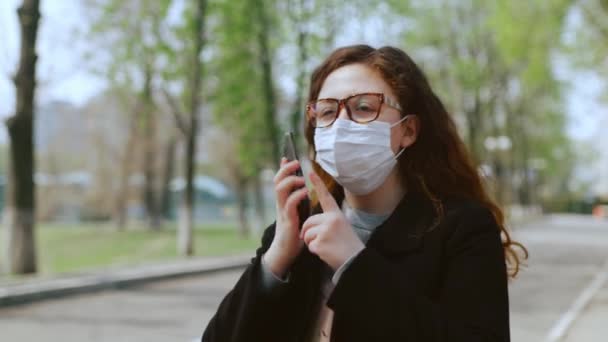 Een jong meisje met een medisch masker gebruikt een smartphone in het park. 4k - Video