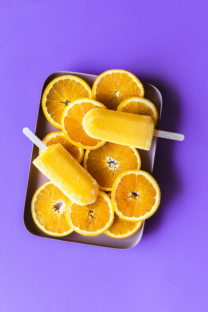 Orange popsicles sorbet and slices of oranges in a tray, top view on a purple background. Delicious homemade orange ice cream - Photo, Image
