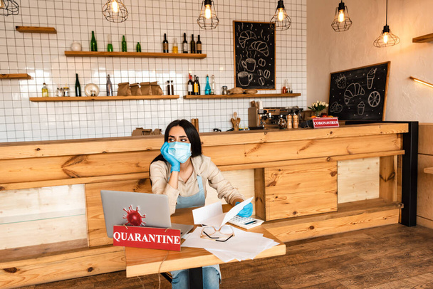 Café proprietário em máscara médica olhando para longe e segurando papel perto de calculadora, documentos, laptop e cartão com letras de quarentena na mesa
 - Foto, Imagem