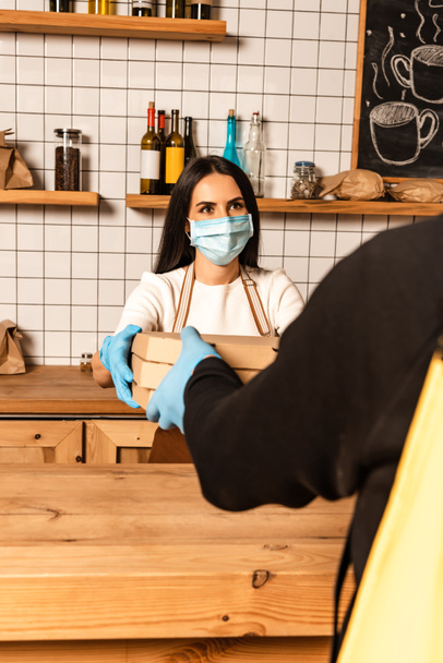 Szelektív fókusz a kávézó tulajdonosa orvosi maszk ad dobozok futár asztal mellett - Fotó, kép