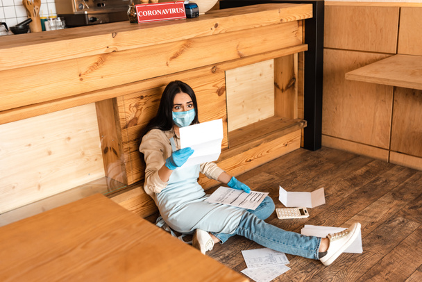 Hoge hoek uitzicht van geschokt cafe eigenaar in medisch masker op zoek naar papier in de buurt van rekenmachine en tafel op de vloer - Foto, afbeelding