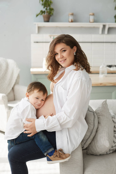 Side view of young pregnant woman with little baby boy posing in home interior. Cute little boy hugging belly of his mother. Cheerful family. Expecting baby - Фото, изображение