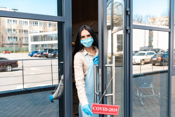 Cafe owner in medical mask near door looking at camera and touching card with covid-2019 lettering on street - Photo, Image