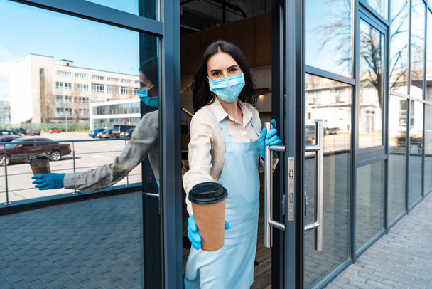 Kávézó tulajdonos orvosi maszk ajtó közelében bemutató eldobható csésze kávét, és nézi a kamera az utcán - Fotó, kép