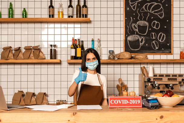 Cafe owner in medical mask showing like sign near table with papers, card with covid-2019 inscription, payment terminal and bowl with fruits - Fotoğraf, Görsel
