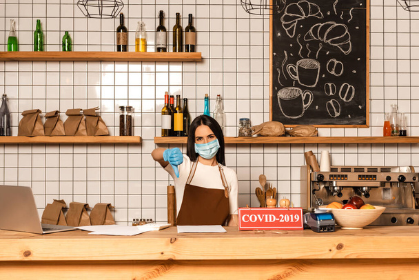Cafe owner in medical mask showing dislike sign near table with papers, card with covid-2019 inscription, payment terminal and bowl with fruits - Foto, Bild