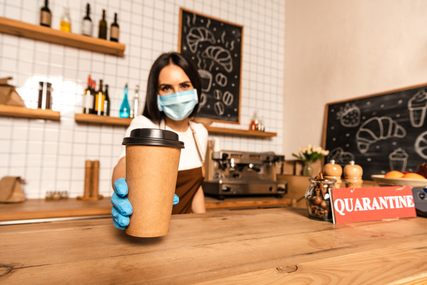 Selective focus of cafe owner in medical mask showing paper cup of coffee near table with card with quarantine inscription - Foto, Bild