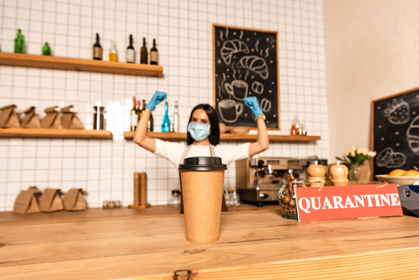 Selective focus of paper cup of coffee near card with quarantine lettering on table and cafe owner in medical mask with hands in air - Photo, Image