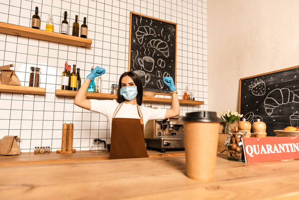 Selective focus of disposable cup of coffee near card with quarantine lettering on table and cafe owner in medical mask with hands in air - Φωτογραφία, εικόνα