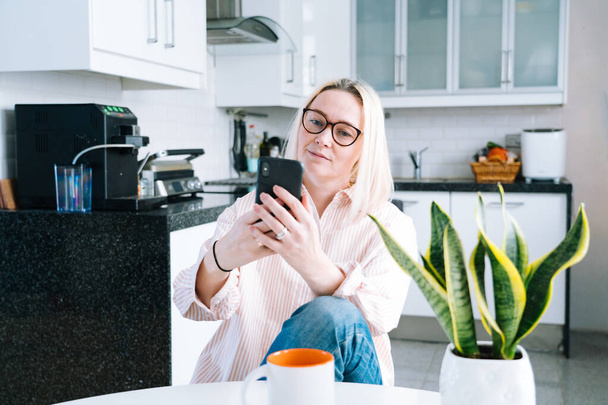Happy girl sitting at home kitchen and holding videocall. Young woman using smartphone for video call with friend or family. Vlogger recording webinar. Woman looking camera and waving greeting hands.  - Fotó, kép