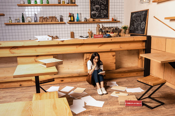 Propriétaire de café choqué regardant calculatrice et assis près de la table, papiers et carte avec lettrage coronavirus sur le sol
  - Photo, image