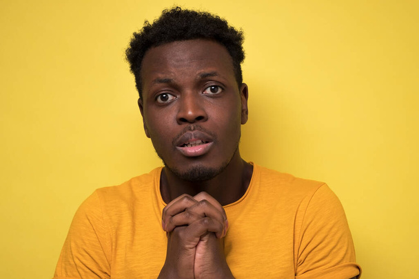 Afrykański amerykański młody człowiek modlący się o najlepsze prosząc o przebaczenie lub cud patrząc przed kamerą, odizolowany na żółtym tle. - Zdjęcie, obraz
